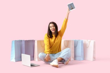 Pembe arka planda tablet bilgisayarı, dizüstü bilgisayarı ve alışveriş çantaları olan genç bir kadın.