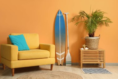 Konforlu koltuk, sörf tahtası, çiçek ve kürek turuncu duvarın yanında.