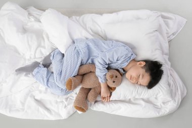 Oyuncak ayı, yastık ve battaniyeli pijamalı şirin Asyalı çocuk beyaz arka planda uyuyor.