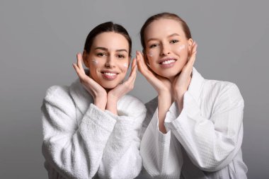 Gri arka planda yüz kremi olan bornozlu güzel kız kardeşler.