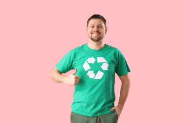 Tişörtünde geri dönüşüm logosu olan mutlu genç adam pembe arka planda baş parmağını kaldırıyor. Ekoloji kavramı