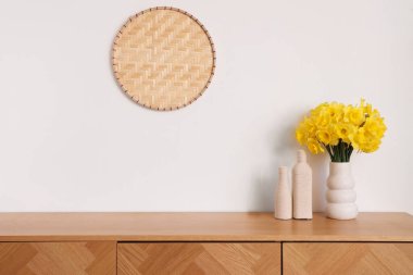 Üzerinde narsis çiçekleri olan vazo, beyaz duvarın yanındaki çekmecelerde, aydınlık odada.