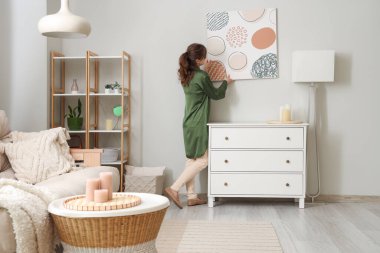 Genç bir kadın şık bir oturma odasında resim asıyor.