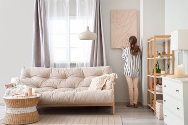 Genç bir kadın şık oturma odasındaki kanepenin yanına resim asıyor.