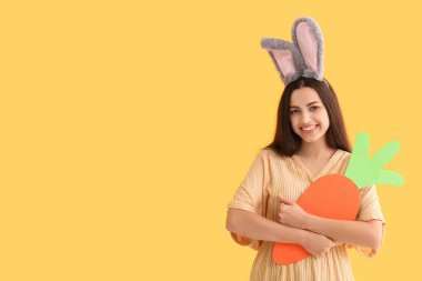 Paskalya tavşanı kulaklı, sarı arka planda kağıt havuç olan güzel, genç bir kadın.