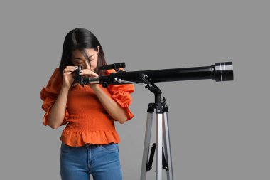 Genç Asyalı kadın gri arka planda teleskop kullanıyor.
