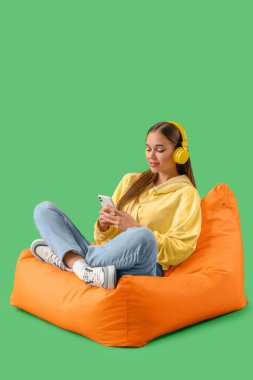 Kulaklıklı genç bir kadın yeşil arka planda cep telefonu kullanıyor.