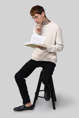 Kitabı olan genç dövmeli adam gri arka planda sandalyede oturuyor.