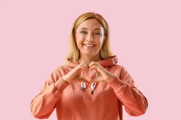 stock image Mature woman in orange hoodie making heart gesture on pink background