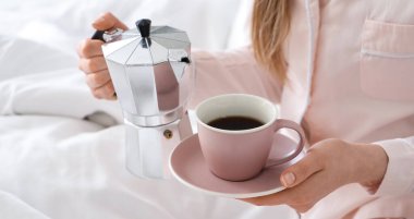 Elinde gayzer kahve makinesi ve yatakta bir fincan espresso tutan bir kadın.