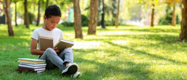 Little African-American boy with books sitting on grass in park