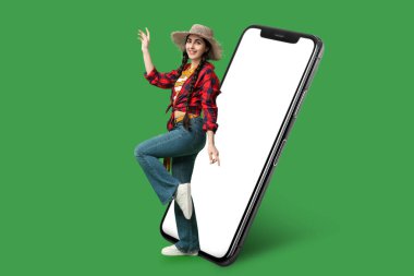 Hasır şapkalı güzel, genç ve mutlu bir kadın ve yeşil arka planda büyük bir akıllı telefon. Festa Junina (Haziran Festivali) kutlaması