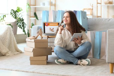 Tableti, kredi kartı, kutuları ve yeni topuklu ayakkabıları olan genç bir kadın evde online alışveriş yapıyor.