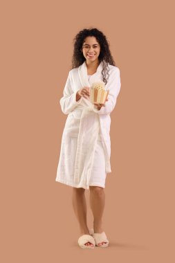 Kahverengi arka planda duş malzemeleri olan bornozlu genç bir Afro-Amerikalı kadın.