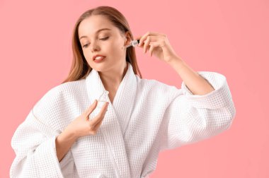 Bornozlu genç bir kadın pembe arka planda serum uyguluyor.