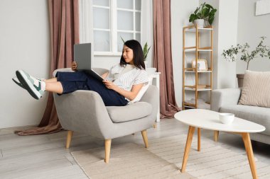 Young Asian woman using laptop in armchair at home