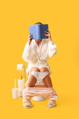 Önlüklü genç kadın sarı arka planda klozet kitabı okuyor.