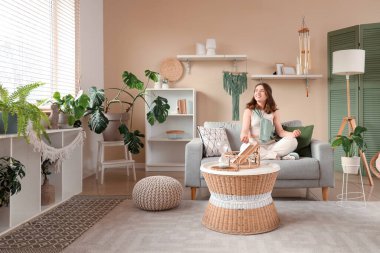 Genç kadın kanepede meditasyon yapıyor ve oturma odasında rüzgar çanı asıyor.