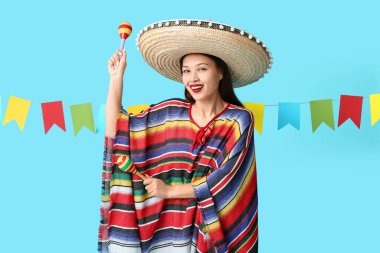 Meksika şapkalı ve pançolu mutlu genç kadın mavi arka planda marakas tutuyor.