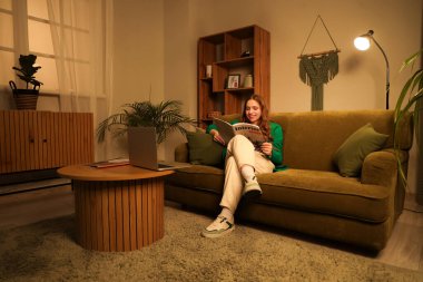 Genç bir kadın akşamları evdeki kanepede dergi okuyor.