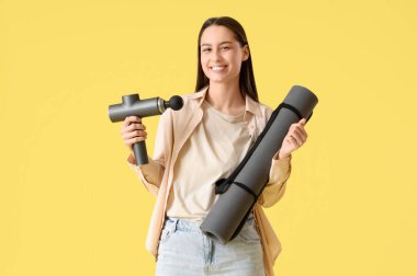 Sarı arka planda vurmalı masaj aleti ve spor paspası olan genç bir kadın.