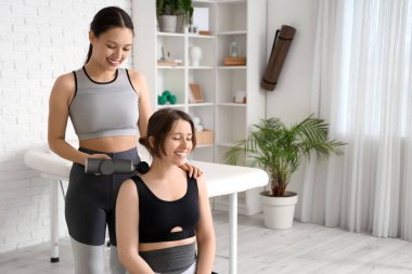 Kadın Asyalı fizyoterapist rehabilitasyon merkezinde genç bir kadının boynuna perküsyon masajı yapıyor.