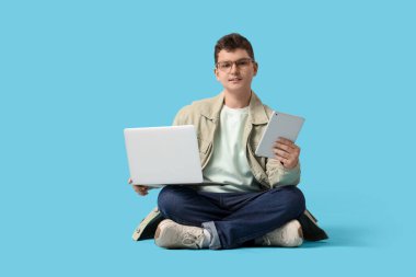 Laptop ve tablet bilgisayarlı genç programcı mavi arka planda oturuyor