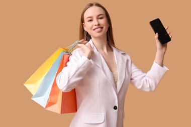 Bej arka planda cep telefonu ve alışveriş çantaları olan genç bir kadın.