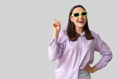 Güneş gözlüklü gülümseyen genç kadın gri arka plandaki bir şeyi işaret ediyor.