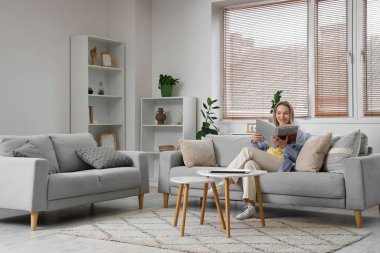 Oturma odasındaki kanepede dergi okuyan genç bir kadın.