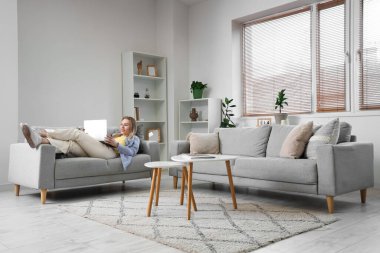 Genç kadın dizüstü bilgisayar kullanıyor ve oturma odasında kanepede yatıyor.