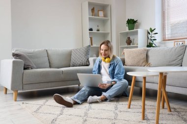 Kulaklıklı genç bir kadın oturma odasında dizüstü bilgisayar kullanıyor.