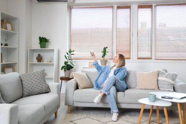 Genç bir kadın oturma odasında klima için uzaktan kumanda kullanıyor.