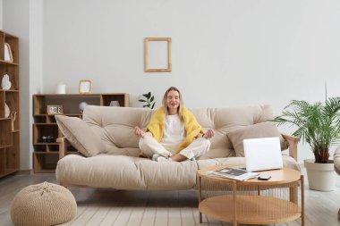 Genç bir kadın şık bir oturma odasında kanepede meditasyon yapıyor.