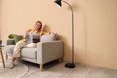 Genç bir kadın dergi okuyor ve bej oturma odasında kanepede oturuyor.