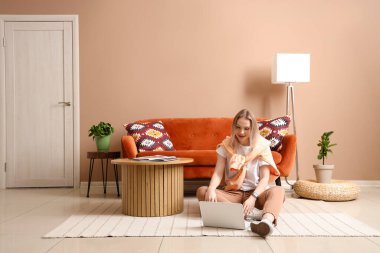 Genç ve güzel bir kadın dizüstü bilgisayar kullanıyor ve bej oturma odasında yerde oturuyor.