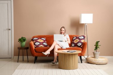 Genç ve güzel bir kadın dizüstü bilgisayar kullanıyor ve bej oturma odasında rahat bir koltukta oturuyor.