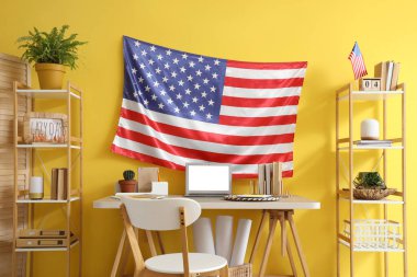 Sarı duvarda işyeri, dizüstü bilgisayar, sandalye ve ABD bayrağı bulunan ofis.