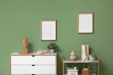 Yeşil duvarın yanında ev bitkileri, şifonyer ve fotoğraf çerçeveleri olan modern raf ünitesi.