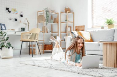 Genç, güzel bir kadın yerde yatıyor ve oturma odasında modern bir laptop kullanıyor.