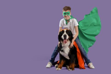 Mor arka planda süper kahraman kostümlü sevimli tüylü köpeği olan mutlu çocuk.