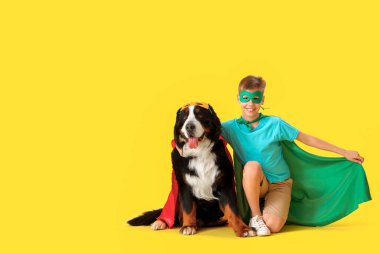 Sarı arka planda oturan sevimli tüylü köpek ve süper kahraman kostümlü çocuk.