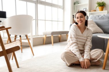 Güzel, genç, mutlu, kulaklıklı bir kadın müzik dinliyor ve evde yerde oturuyor.