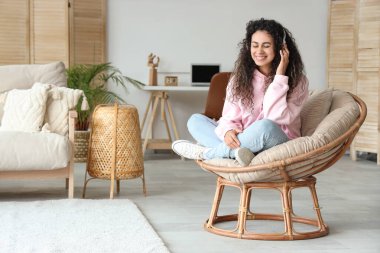 Kulaklıklı genç Afro-Amerikalı kadın evdeki koltukta dinleniyor.