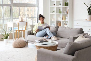 Young African-American woman reading magazine on sofa at home