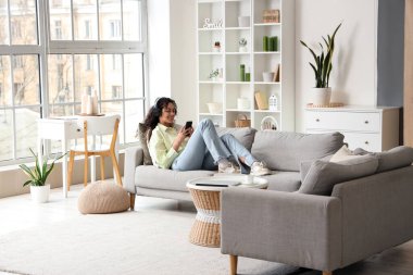 Kulaklıklı genç Afro-Amerikalı kadın evdeki koltukta cep telefonu kullanıyor.