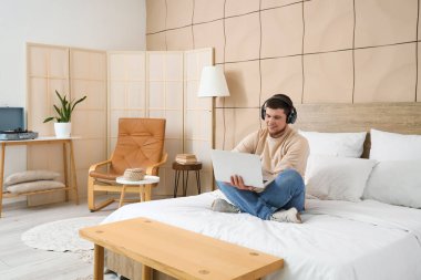 Kulaklıklı genç sakallı adam yatak odasında dizüstü bilgisayar kullanıyor.