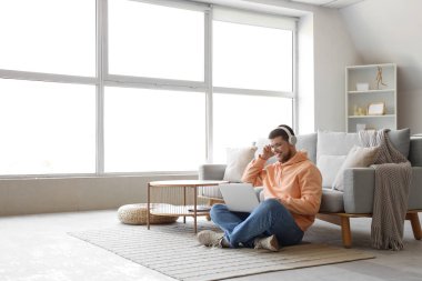 Kulaklıklı, dizüstü bilgisayarlı, evde müzik dinleyen genç sakallı adam.