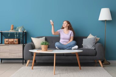 Mavi duvarın yanındaki kanepede oturan klimalı uzaktan kumandalı gülümseyen genç kadın.