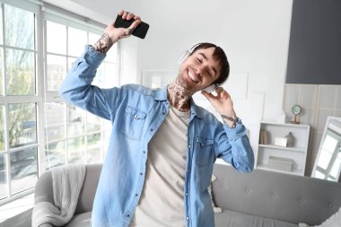 Kulaklıklı, cep telefonlu, evde müzik dinleyen dövmeli genç bir adam.